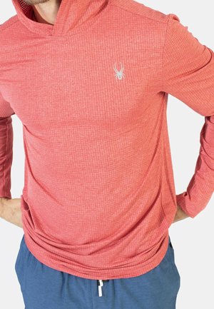Man draagt een rood gestructureerd hoodieshirt met lange mouwen en een wit spinlogo op de borst en blauwe broek met aantrekkoord, handen in de heupen.