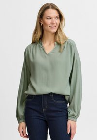 Blusa verde a maniche lunghe con scollatura a V e tessuto testurizzato, dotata di maniche arricciate e vestibilità comoda, abbinata a jeans scuri.