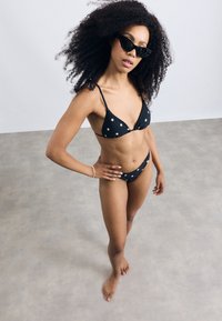 Set da bikini nero con motivo a pois, composto da reggiseno triangolare e slip coordinato. Modella con occhiali da sole neri oversize. Ambientazione interna.