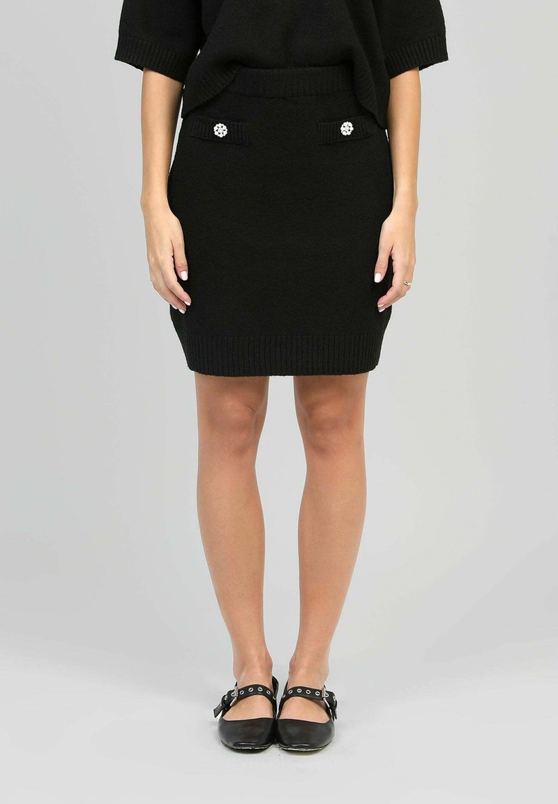 Jupe crayon noire en tricot avec une taille côtelée, dotée de deux poches ornées de boutons décoratifs en argent. Associée à des ballerines noires.