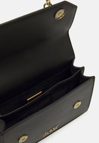Černá kožená kabelka se zlatými magnetickými knoflíky, otevřená, aby ukázala dvě vnitřní přihrádky a zlatou značkovou plaque uvnitř.