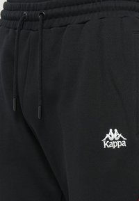 Pantalones deportivos negros con cintura elástica y cordón. Presentan un pequeño logo bordado en blanco en el muslo. El material parece suave y texturizado.