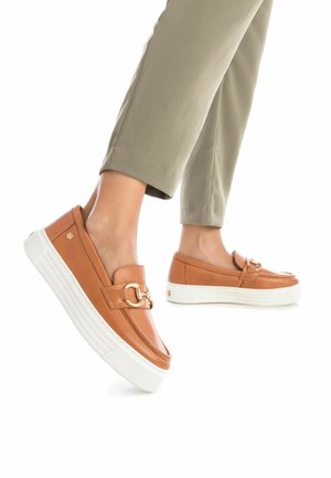 Scarpe slip-on in pelle color tan con un accento circolare dorato, suola piatta bianca e superficie liscia, abbinate a pantaloni verde oliva chiaro.