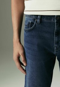 Tmavomodré džínsy s úzkym strihom, s prednou vreckom, viditeľným šitím a kovovým zapínaním na gombík. Mäkká textúra látky.