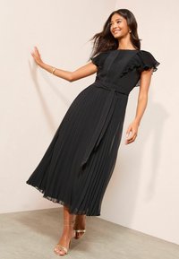 Must, voldikleit, millel on volangidega varrukad ja vöökumm; kleidi ülaosa on tekstuuriga ning kangas voogab ja on läbipaistev. Kuldset värvi sandaalid.