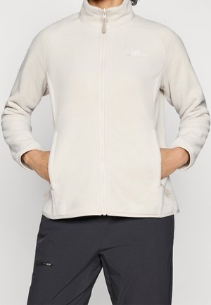 Henkilö pukeutuneena vaalean beigeen täysvetoketjulliseen fleecetakkiin, kädet sivutaskuissa ja tummat housut yksiväristä taustaa vasten.