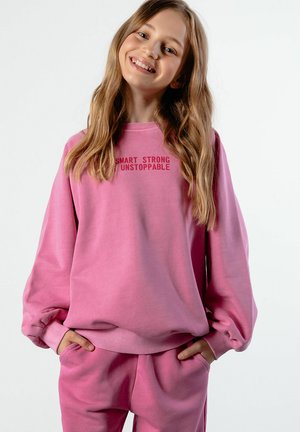 Uśmiechnięta dziewczyna z długimi włosami, ubrana w różową bluzę i spodnie, na bluzie napis "Smart Strong Unstoppable,", ręce w kieszeniach.