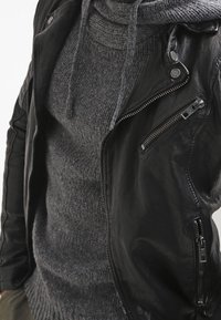 Giacca da moto in pelle nera con accessori in argento, tasche con zip e texture a coste. Indossata sopra un maglione grigio a maglia con collo a cascata.