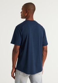 T-shirt in cotone blu navy con maniche corte, colletto a giro e un orlo posteriore più lungo. Texture morbida, design minimalista, senza motivi o accessori visibili.