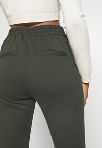 Tumman oliivinvihreät joogahousut, joissa on joustava vyötärö, kaksi takataskua ja sileä, pehmeä kangas.