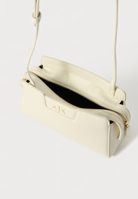 Krémová crossbody taška s hladkým povrchem, zlatým zipem a embosovaným logem. Obsahuje zipovou přihrádku a nastavitelný popruh.