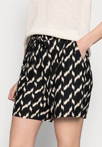 Shorts noirs avec un motif en zigzag ivoire, dotés d'une taille élastique avec cordon de serrage et de poches latérales. Fabriqués en tissu léger.