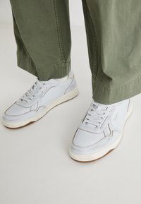 Baskets bleu clair avec des lacets blancs et des accents marron. Design basse avec des détails perforés et une semelle en caoutchouc texturée. Pantalons vert olive.
