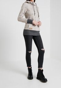 Kvinna klädd i en beige hoodie med grå detaljer, trasiga svarta skinny jeans och svarta kängor med snörning, stående mot en enfärgad bakgrund.