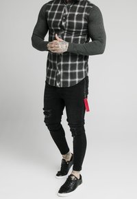 Camisa de cuadros en negro y gris con botones y mangas largas, combinada con jeans skinny negros rasgados y zapatillas negras con detalles dorados.