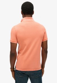 Korte mouwen, koraal polo met een gestructureerde afwerking en kraag, gecombineerd met donkerblauwe jeans, vanaf de achterkant bekeken.