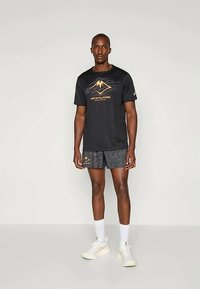 Černé tričko ASICS s oranžovou grafikou hory, spárované s šedými vzorovanými šortkami. Model má na sobě bílé boty se světlými podrážkami a bílé ponožky.