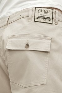 Bežīgas kokvilnas bikses ar aizmugurējo kabatu, kas aprīkota ar sudraba pogu. Uz jostas ir izšūta "GUESS" etiķete ar melnu un baltu detaļu.