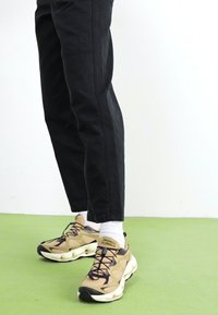 Zapatillas beige con acentos negros, cordones y una suela acolchada, combinadas con pantalones negros y calcetines blancos, posicionadas sobre una superficie verde.