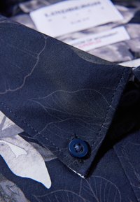 Dettaglio di una camicia floreale blu navy con tessuto strutturato, motivo floreale chiaro a contrasto e un bottone blu sul colletto.