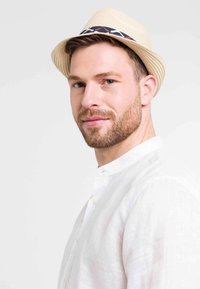 Bež slamnati fedora s plavo-belo geometrijsko trakom, nošen uz belu košulju sa stajaćim kragnom, od gladkog materijala.