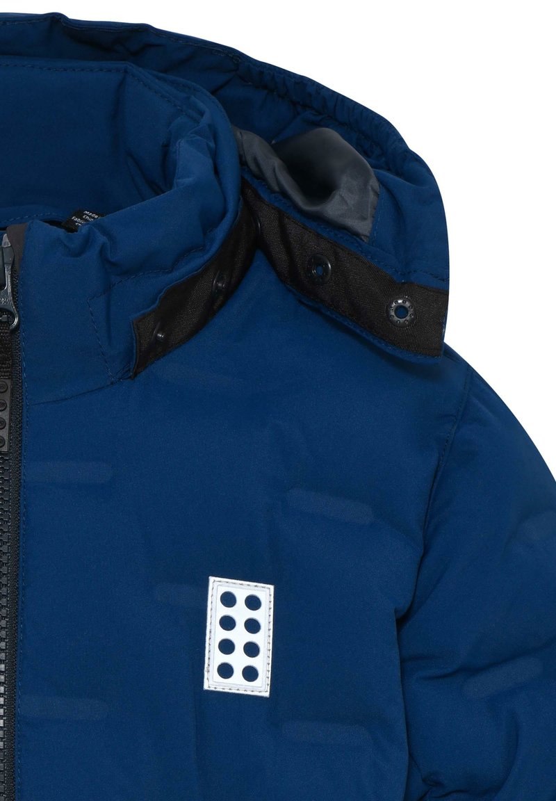 Veste matelassée bleu marine avec fermeture éclair à l'avant, col intérieur noir et insigne blanc avec cinq découpes circulaires sur la poitrine gauche.