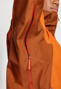 Braune und orangefarbene Jacke mit glatter Textur, ausgestattet mit einem diagonalen Reißverschluss in Rot und orangefarbenen Akzenten. Zusätzliche Taschen vorhanden.