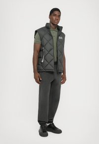 Black puffer vest with diamond pattern, no sleeves, zippered pockets; worn over a green polo and paired with dark pants and black shoes.