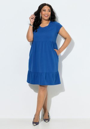 Mujer sonriendo y posando con la mano en el bolsillo, vistiendo un vestido azul de manga corta y tacones de punta, sobre fondo blanco.