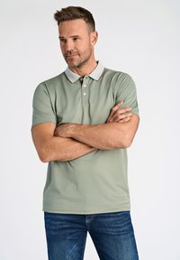 Pikétröja i ljusgrön med grå krage och bruna accenter; kortärmad; slät tygstruktur; bärs med mörkblå jeans.