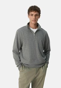 Pulover gri cu dungi, cu guler cu fermoar pe jumătate, fabricat dintr-un material moale și texturat, asortat cu pantaloni verzi deschis.