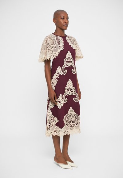 Vestido bordô com detalhes em renda creme, apresentando mangas curtas e uma bainha com bordas onduladas. A renda tem padrões florais e espirais intrincados.