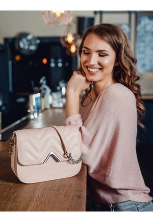 Cremfarbener Lederhandtasche mit einem strukturierten, gewellten Design, metallischen Akzenten und einem Kettenriemen, der auf einer Holzbar-Theke ruht.