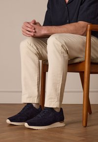Sneakers in suede navy con suole beige, caratterizzati da una superficie testurizzata e lacci piatti, indossati con pantaloni color crema mentre si è seduti su una sedia di legno.