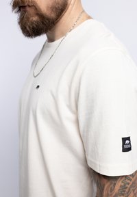 T-shirt in cotone beige con maniche corte, caratterizzato da un piccolo logo di un orso nero sul petto e un'etichetta nera sulla manica sinistra.