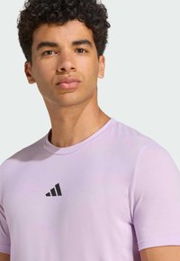 Camiseta atlética de color púrpura claro con cuello redondo, mangas cortas y un logo negro de Adidas en el pecho. Textura de tela suave y ajustada.