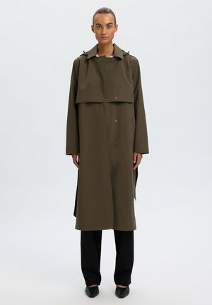 Femme debout, face à l'avant, portant un long trench-coat vert olive, un pantalon noir et des chaussures plates noires sur un fond clair uni.