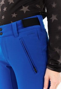 Close-up van blauwe broek met een zijritszak en verstelbare zwarte klittenbandsluiting, gedragen met een zwart top met grijze sterrenpatronen.