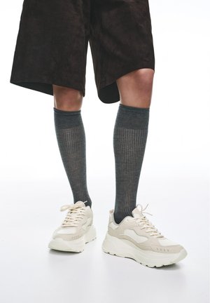 Fekete rövidnadrágot, bordázott sötétszürke térdzoknit és vastag, törtfehér cipőfűzős tornacipőt viselő lábak fehér háttér előtt.