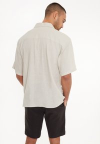 Kurzärmliges, hellbeiges Leinenhemd mit entspannter Passform und Rückenpasse. Kombiniert mit schwarzen Shorts, die einen legeren Stil präsentieren.