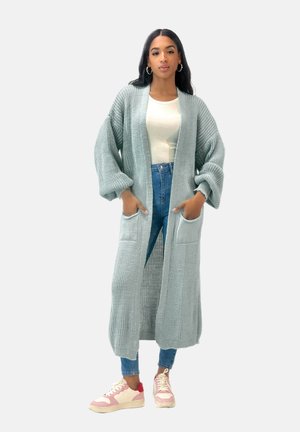 Langer, hellblauer Strickcardigan mit übergroßen Ärmeln, Fronttaschen und einem strukturierten Muster. Getragen über einem weißen Oberteil und blauen Jeans.