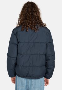 Persona con capelli lunghi e ricci che indossa una giacca a vento blu scuro e jeans a fantasia blu chiaro, in piedi e girato di spalle su uno sfondo bianco.