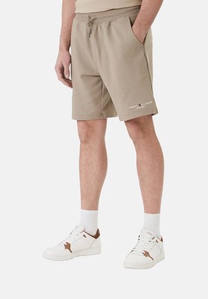 Hombre vistiendo pantalones cortos beige de Tommy Hilfiger con bolsillos, calcetines blancos y zapatillas blancas con detalles marrones sobre un fondo claro.