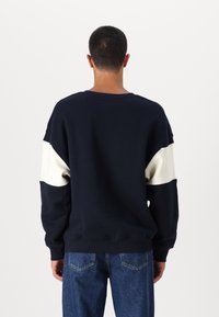 Marine sweatshirt met een crème streep, ronde hals en geribbelde mouwen. Zachte katoenmix, ontspannen pasvorm en minimale stiksels aan de achterkant.
