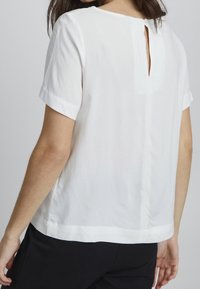 Vit kortärmad blus med en nyckelhålsöppning bak, sidofickor och mjukt tyg. Enkel design med en elegant finish.