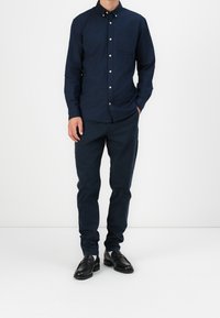 Camicia blu navy con bottoni e taschino sul petto, abbinata a pantaloni blu navy scuro e mocassini neri in pelle, posata davanti a uno sfondo bianco.