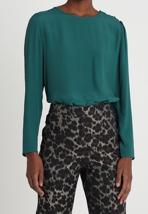 Modelo vistiendo una blusa de manga larga en color azul verdoso con detalles en los hombros, combinada con pantalones de estampado de leopardo de cintura alta, con las manos descansando a los lados.