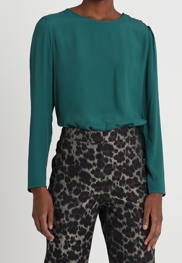 Modelka v tyrkysové blůze s dlouhými rukávy a detaily na ramenou, spárované s kalhotami s vysokým pasem a leopardím potiskem, ruce položené na bokách.
