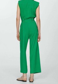 Combinaison verte sans manches avec une taille cintrée et des jambes larges. Caractérisée par des coutures au dos et des boutons. Associée à des sandales argentées à brides.