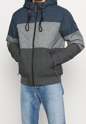 Homme portant une veste à capuche avec des panneaux bleu, gris et gris foncé et un jean bleu clair, mains dans les poches de la veste.